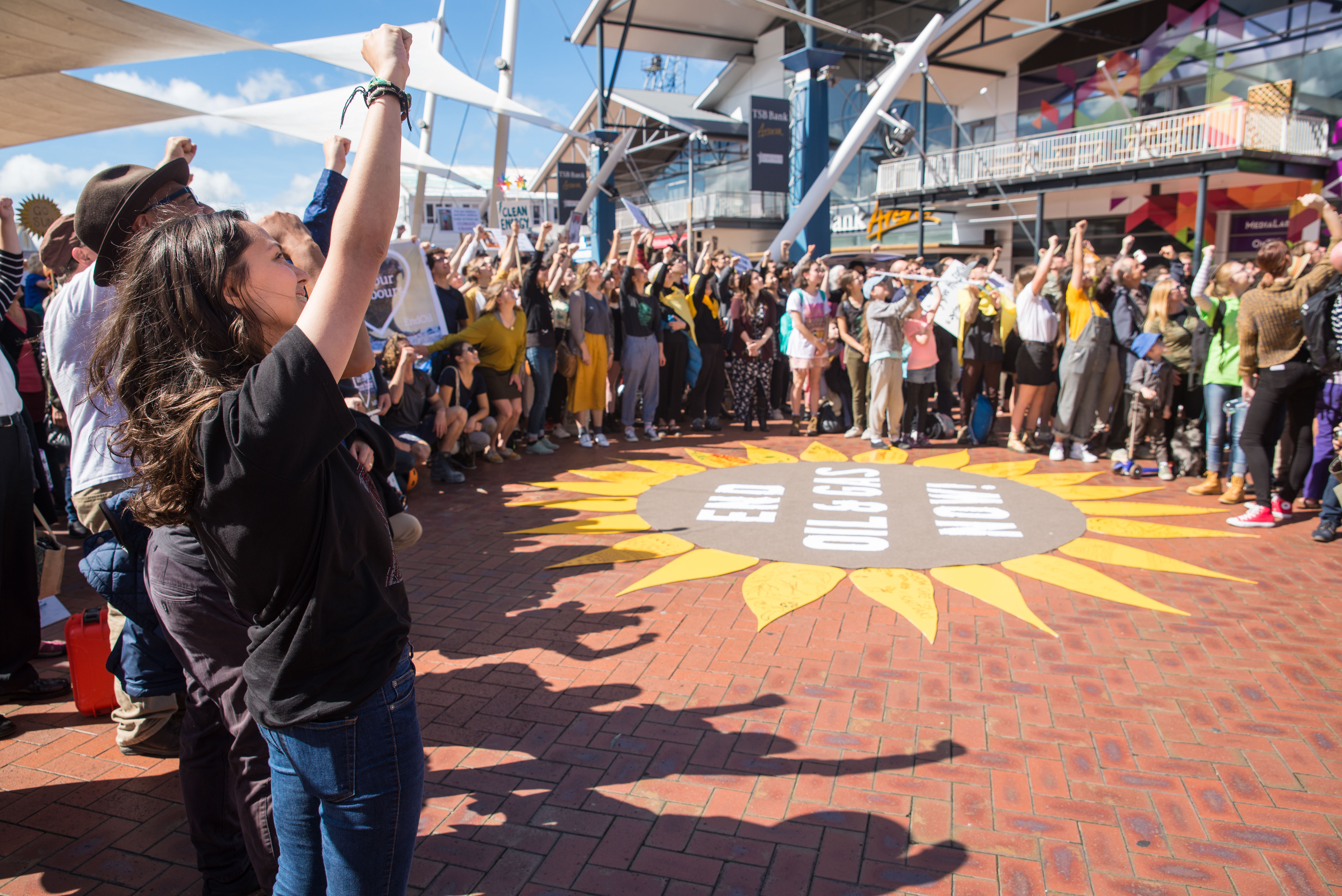 People standing fists raised in a crowd around giant sunflower that reads "End Oil and Gas Now!"